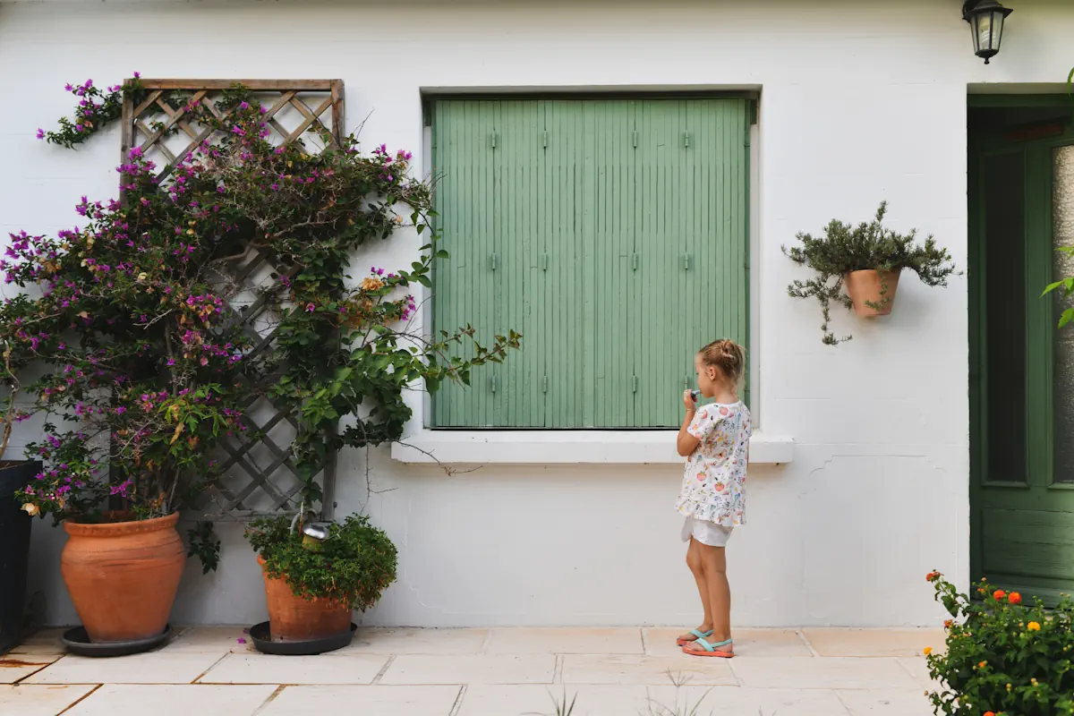 Une petite fille devant un volet vert sauge avec des plantes devant
